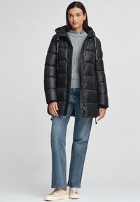 Svart vadderad jacka med huva, med horisontell sömnad, dragkedja och dragsnören, bärs över en grå stickad tröja och blå jeans.