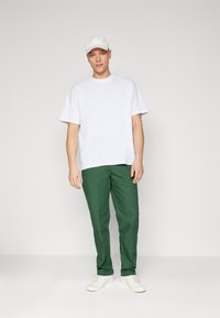 Tricou alb din bumbac, pantaloni verzi din bumbac și adidași albi. Modelul stă în fața unui fundal simplu, purtând o șapcă albă.
