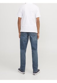 Persona de pie de espaldas, vistiendo una camiseta blanca de manga corta, jeans azules y zapatillas en blanco y negro, sobre un fondo sencillo.