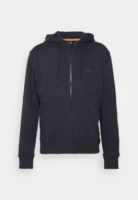Sudadera con cremallera de color azul marino, hecha de una mezcla de algodón, que presenta una capucha con cordón, un forro naranja contrastante y un pequeño parche con el logo en el pecho.
