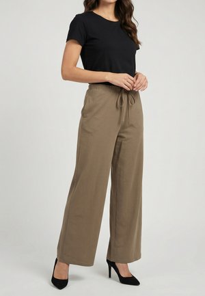 Mujer con camiseta negra de manga corta, pantalones anchos beige con cintura con cordón y zapatos negros de tacón alto, de pie frente a un fondo neutro.