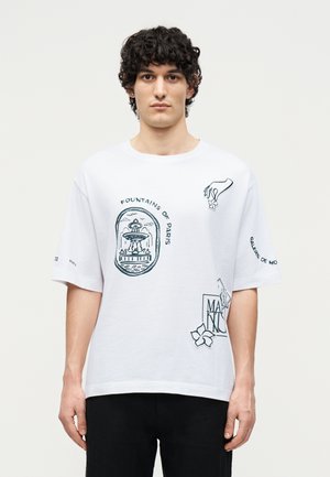 Homme aux cheveux bouclés portant un t-shirt blanc oversize avec des illustrations noires de fontaine, de fleur et de main, et un pantalon noir sur un fond uni.