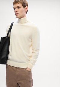 Pulover cu guler înalt de culoare crem, dintr-un material tricotat moale, asortat cu pantaloni maronii și o geantă de umăr neagră. Design simplu, ajustat, cu manșete zimțate.