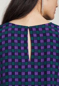 Vestuário de manga curta com um padrão xadrez roxo, verde e preto. Apresenta uma abertura em forma de lágrima na parte de trás do decote. Tecido suave e texturizado.