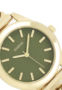 Montre dorée avec un boîtier rond et un cadran texturé vert. Comprend des index et aiguilles dorés, avec un bracelet métallique classique.