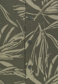 Olivenfarbener Hemdenstoff mit einem cremefarbenen, abstrakten Blattmuster, an dem vier runde graue Knöpfe sichtbar entlang der Vorderseite angebracht sind.