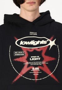 Svart hettegenser med grafisk trykk som har rød og hvit tekst. Merkes noen detaljer som en stjernestråle-designet og ulike logoer og mål.