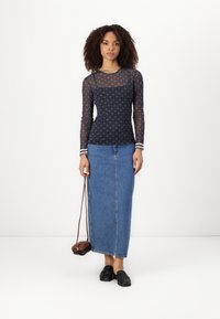 Lange mouwen marineblauw mesh top met sterrenpatroon, gecombineerd met een lichtblauwe denim maxi rok. Model draagt een bruine schoudertas en zwarte schoenen.