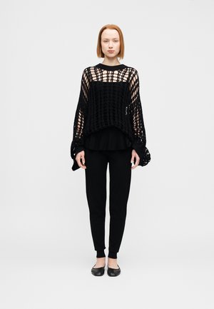 Pull noir à maille ajourée avec manches longues et ourlet côtelé, porté par-dessus un haut noir uni, associé à un pantalon noir slim et des ballerines noires.