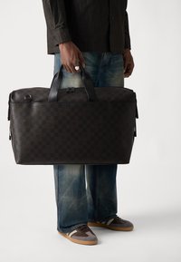 Dark brown leather duffel bag with a checkerboard pattern, featuring dual handles and a zipper. Paired with jeans and sneakers.