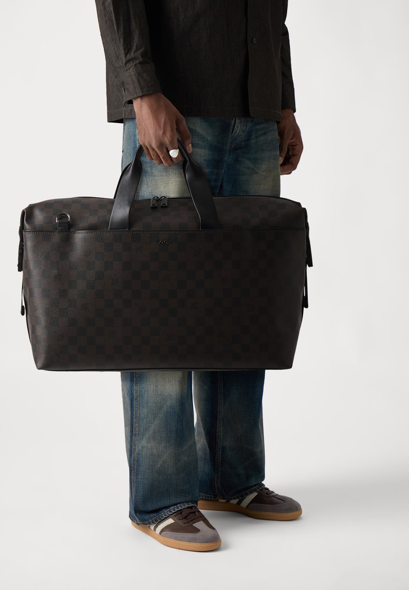 Dark brown leather duffel bag with a checkerboard pattern, featuring dual handles and a zipper. Paired with jeans and sneakers.