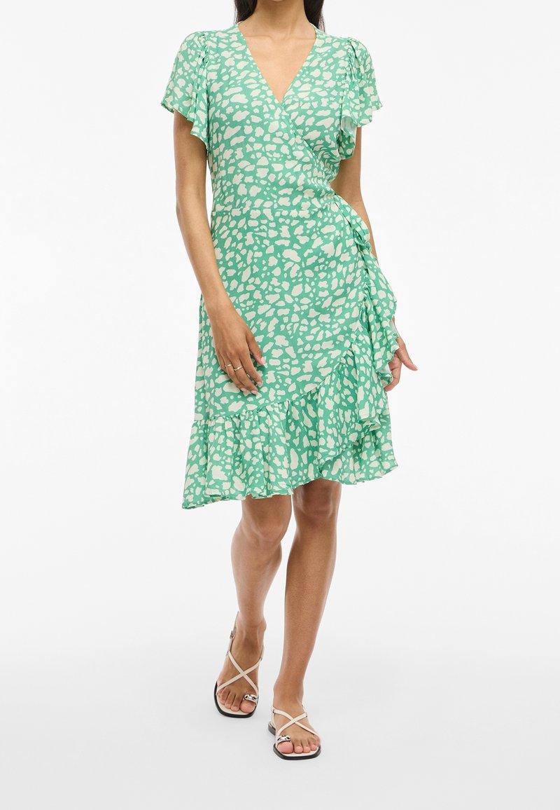 Robe portefeuille verte avec un motif abstrait blanc, manches évasées et ourlet froncé. Tissu léger avec un décolleté en V.