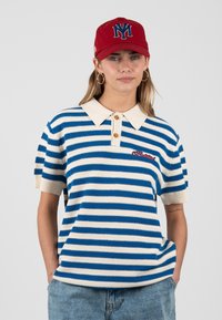 Mujer que lleva una camisa polo de manga corta a rayas azules y blancas, gorra de béisbol roja con el logo "NY", pendientes de aro dorados y pantalones vaqueros, con las manos en los bolsillos.