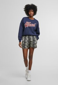 Námořnická mikina s červeným a bílým logem, spárovaná se zelenými šortkami s hadím vzorem. Model má na sobě bílé tenisky a bílé režné ponožky.