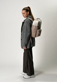 Zaino in tessuto grigio con un pannello superiore crema, due tasche laterali e accenti beige. Modello che indossa un blazer grigio e pantaloni scuri.