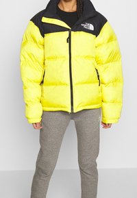 Żółta kurtka puchowa z czarnym kołnierzem i zamkiem z przodu. Posiada duże pikowane sekcje oraz logo na lewym ramieniu. Noszona z szarymi spodniami.