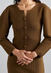 Brun ribbad stickad cardigan med rund halsringning, som har fem knappar klädda med tyg framtill och långa ärmar.