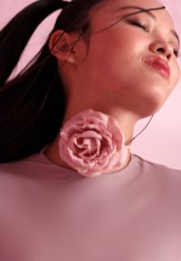 Chokerhalsband med en stor, texturerad ljusrosa ros, fäst vid en mjuk, figurnära ljusgrå topp som framhäver linjen på halsen.