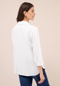 Femme aux cheveux foncés de longueur moyenne, portant une blouse blanche à manches longues et un jean bleu, se tenant de profil devant un fond neutre.