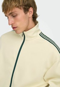 Jeune homme aux cheveux blonds courts portant une veste crème à fermeture éclair avec des rayures vert foncé sur les épaules, regardant vers le bas.