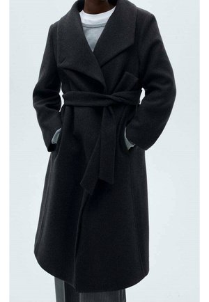 Ciemnoszary wełniany płaszcz z paskiem i szerokimi klapami, noszony na szarym swetrze i białej koszuli, dłonie w kieszeniach, pokazany od połowy uda do ramion.