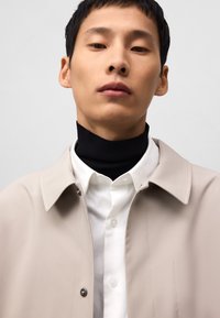 Beige buitenjack over een zwart coltrui en wit shirt, met een klassieke kraag en drukknoopsluiting. Glad van structuur en minimalistisch design.