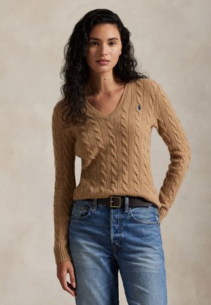 Frau mit langen lockigen dunklen Haaren, die einen beigen kabelgestrickten Pullover mit V-Ausschnitt und blaue Jeans trägt, steht vor einem schlichten beigen Hintergrund.
