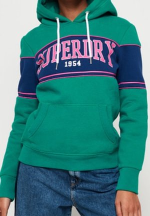 Zielono-granatowa bluza z kapturem z różowym napisem "Superdry 1954", noszona z niebieskimi dżinsami, z kieszenią z przodu i białymi sznurkami.