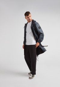 Marinblå vattentät jacka med huva, tryckknappar och patchdetalj, bärs över en vit t-shirt, ihop med svarta byxor och sneakers.