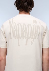 Beige cotton t-shirt with short sleeves, featuring a large grey logo text "Napapijri" printed on the back. Smooth texture and classic fit.
