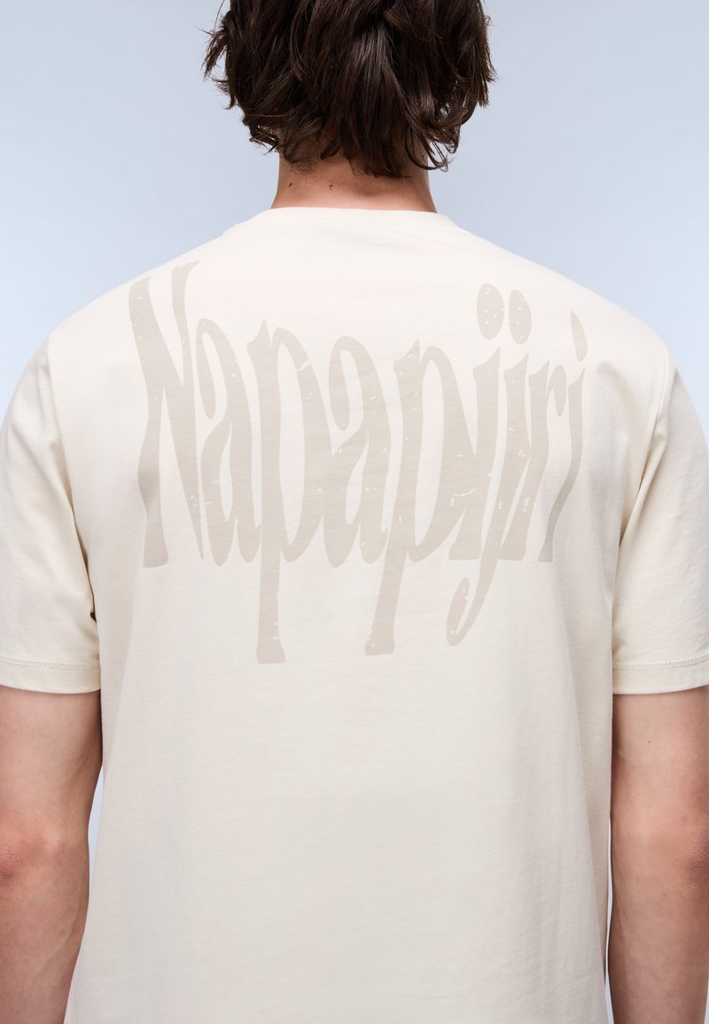 T-shirt en coton beige à manches courtes, avec un grand logo textuel gris "Napapijri" imprimé au dos. Texture lisse et coupe classique.