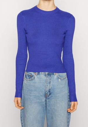 Personne portant un pull violet ajusté à manches longues en côte, rentré dans un jean taille haute bleu clair.
