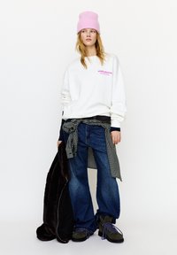 White sweatshirt with pink print, blue jeans, layered checkered shirt tied at the waist, brown fur coat, pink beanie, and gray boots.