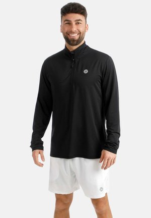 Lächelnder Mann, der einen schwarzen Langarm-Halbzipper-Pullover und weiße Shorts trägt, vor einem schlichten hellen Hintergrund stehend.