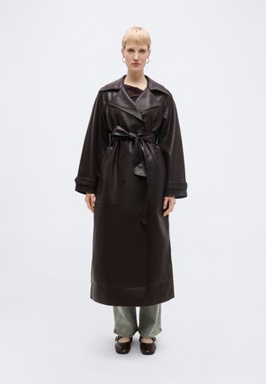 Γυναίκα με κοντά ξανθά μαλλιά που φοράει μακρύ, σκούρο καφέ δερμάτινο παλτό τρανς δεμένο στη μέση, γκρι παντελόνι φαρδιάς γραμμής και σκούρα καφέ παπούτσια σε λευκό φόντο.