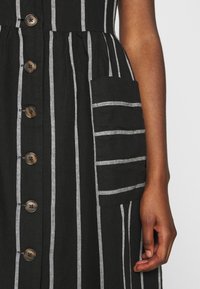 Robe à rayures noires avec des rayures grises verticales, boutons sur le devant, et une poche latérale avec des accents de rayures horizontales.