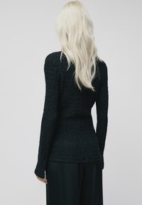Maglietta nera a maniche lunghe realizzata in tessuto testurizzato con un design aderente e maniche affusolate, abbinata a pantaloni scuri. È visibile un sottile motivo a onde.