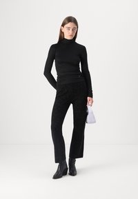 Haut noir à manches longues et col roulé, associé à un pantalon noir texturé avec des éclats argentés. Tenant un petit sac à main blanc, portant des bottines noires.