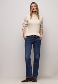 Blondýna v krémovém svetru, modrých džínách a hnědých botách stojí s rukama v kapsách u světle šedé zdi.