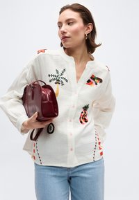 Femme portant une chemise blanche avec une broderie colorée de fruits et d'arbres, tenant un sac à main en cuir rouge, et portant un jean bleu clair.