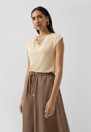 Vrouw met lang donker haar draagt een beige mouwloos topje met gedrapeerde halslijn, een bruine hoge taille rok met trekkoord en gouden oorringen.