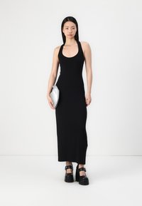 Svart, ermeløs maxikjole med rund hals, tettsittende silhuett, glatt tekstur. Modellen holder en sølvfarget clutch og har på seg kraftige svarte hæler.
