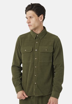 Uomo che indossa una camicia di velluto a coste verde oliva con due tasche sul petto, che guarda a sinistra contro uno sfondo semplice.