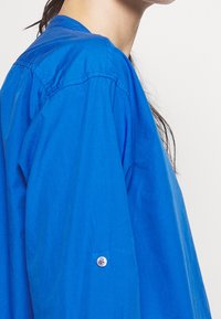 Helles blaues Hemd mit spitzem Kragen, langen Ärmeln und abgerundetem Saum. Der Stoff wirkt leicht und hat eine glatte Textur.