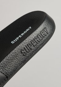 Zwarte textuur slide sandaal met een verhoogd "SUPERDRY" logo in het wit. Het zachte leren materiaal heeft een smooth afwerking en een gevormde vorm.