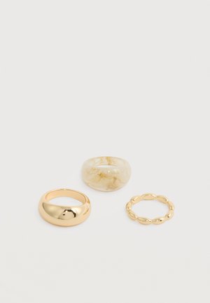 Trois bagues sur une surface blanche : une en or brillant en forme de dôme, une grosse marbrée crème, et une fine en or avec des perles arrondies.