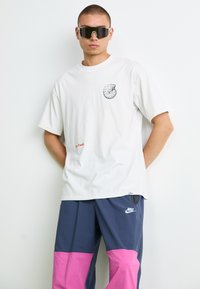 Homme portant un t-shirt blanc surdimensionné avec un graphique circulaire noir et un pantalon Nike bleu-rose, se tenant les mains derrière le dos contre un mur blanc.