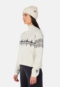 Light-colored knitted sweater with black geometric pattern, mock neck, and ribbed cuffs. Paired with a matching ribbed cream beanie.
