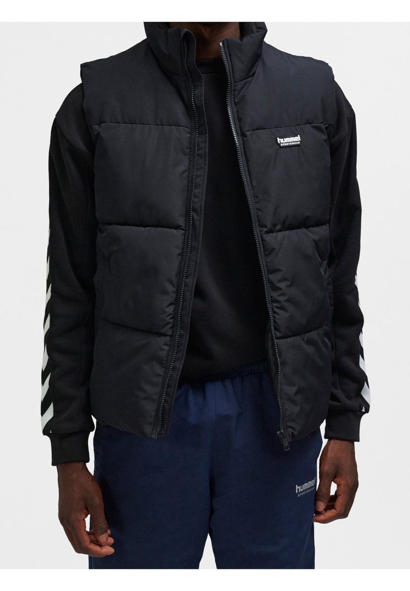 Homme portant une doudoune noire sans manches sur un haut noir à manches longues avec des rayures en chevrons et un pantalon bleu marine avec le logo Hummel.