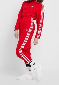 Röd träningsoverall med vita ränder, dragkedja vid kragen och logotyp på bröstet och byxorna. Kombinerad med vita sneakers och randiga strumpor.